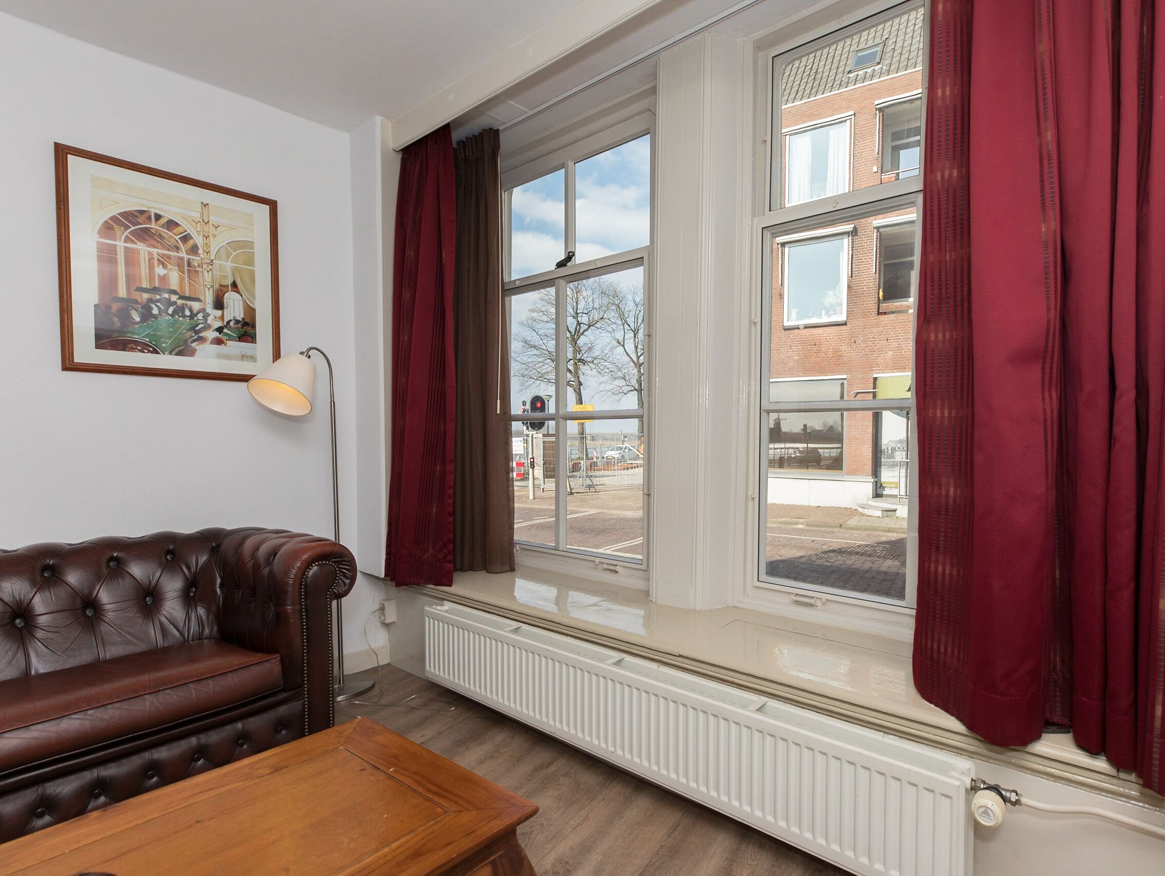 Una habitación con sofá de cuero marrón, una mesa de centro de madera, una lámpara de pie y una ventana con cortinas rojas.