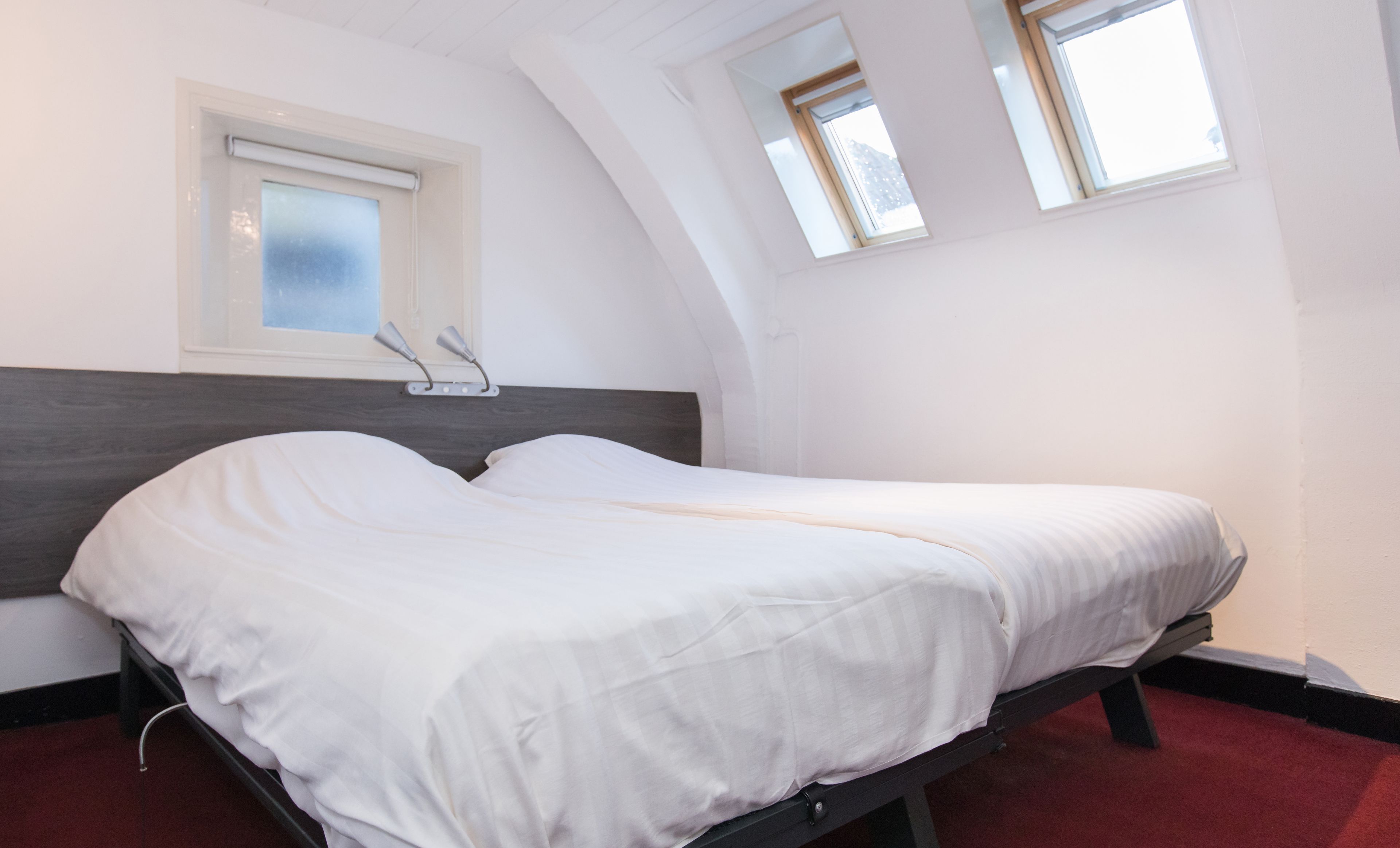 Un letto singolo in una stanza con soffitto spiovente, una finestra con vetro smerigliato e un muro bianco liscio.