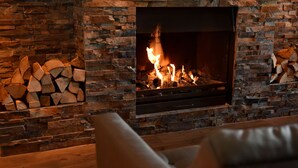 A stone fireplace with a fire burning inside, surrounded by stacked firewood.