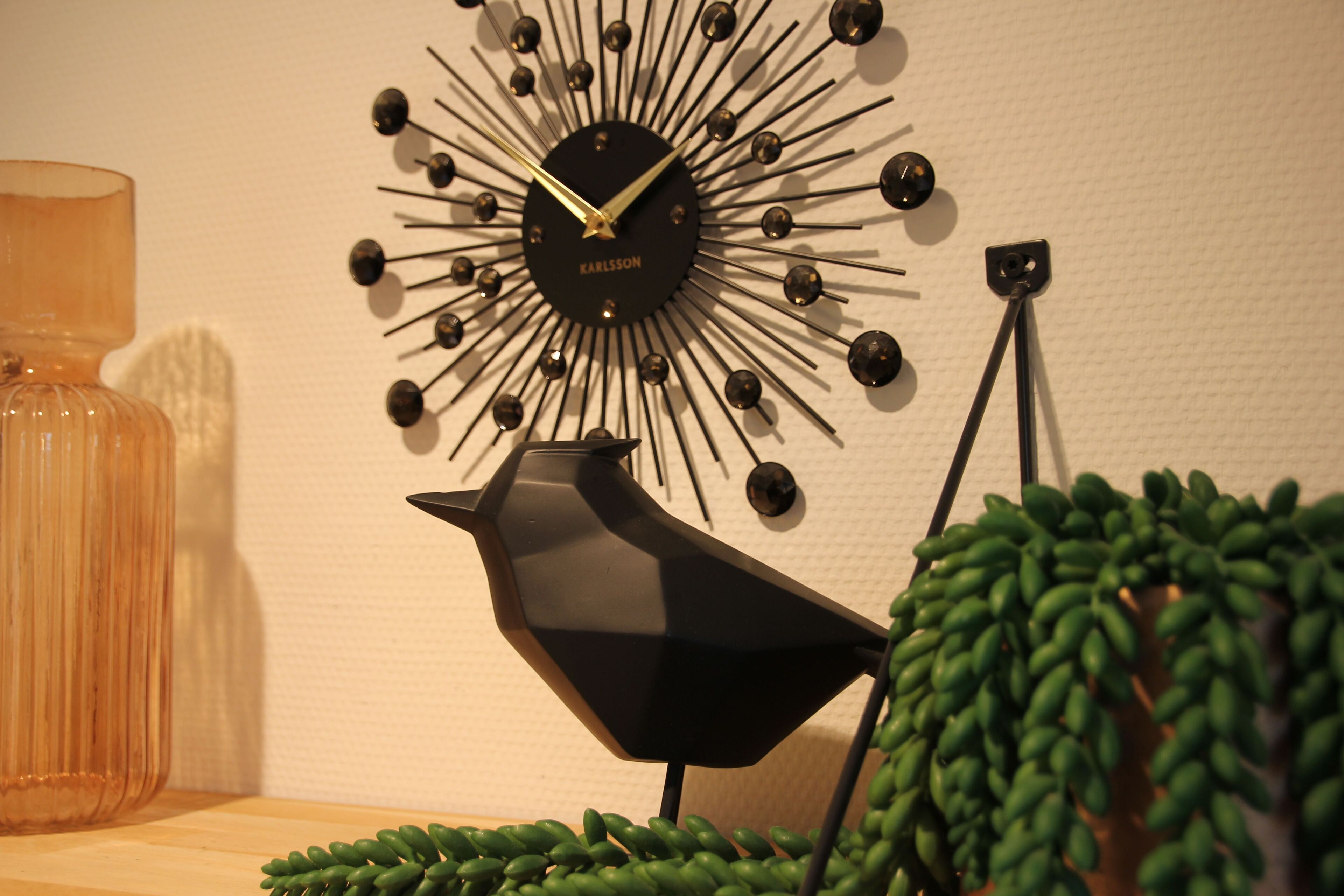 Een wandklok met een zonnewiel ontwerp, een zwarte lamp in de vorm van een vogel, en een groene plant.