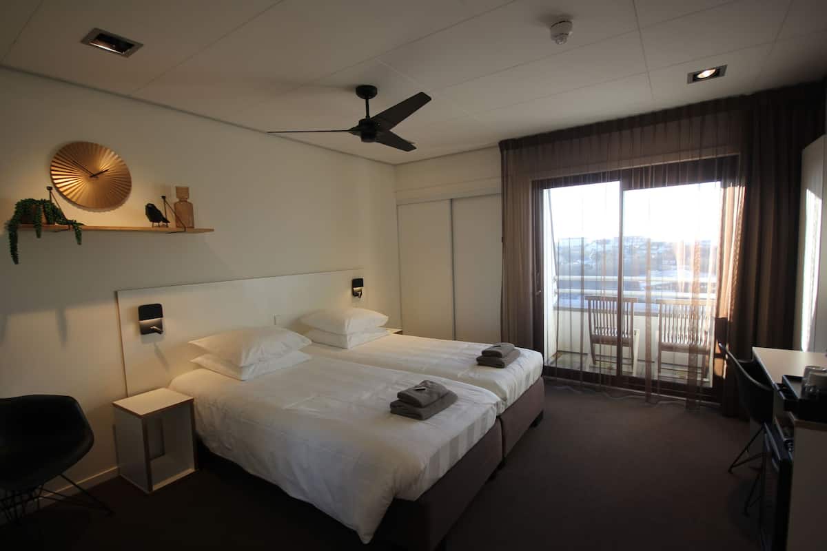 A hotel room with two beds, a desk, a chair, a ceiling fan, a window with curtains, and a view of the city.
