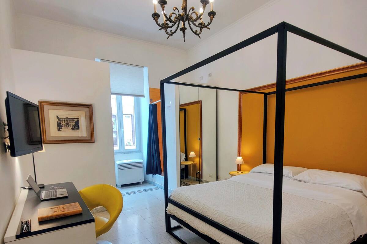A four-poster bed, a desk with a laptop, a chair, a television, and a framed picture on the wall.