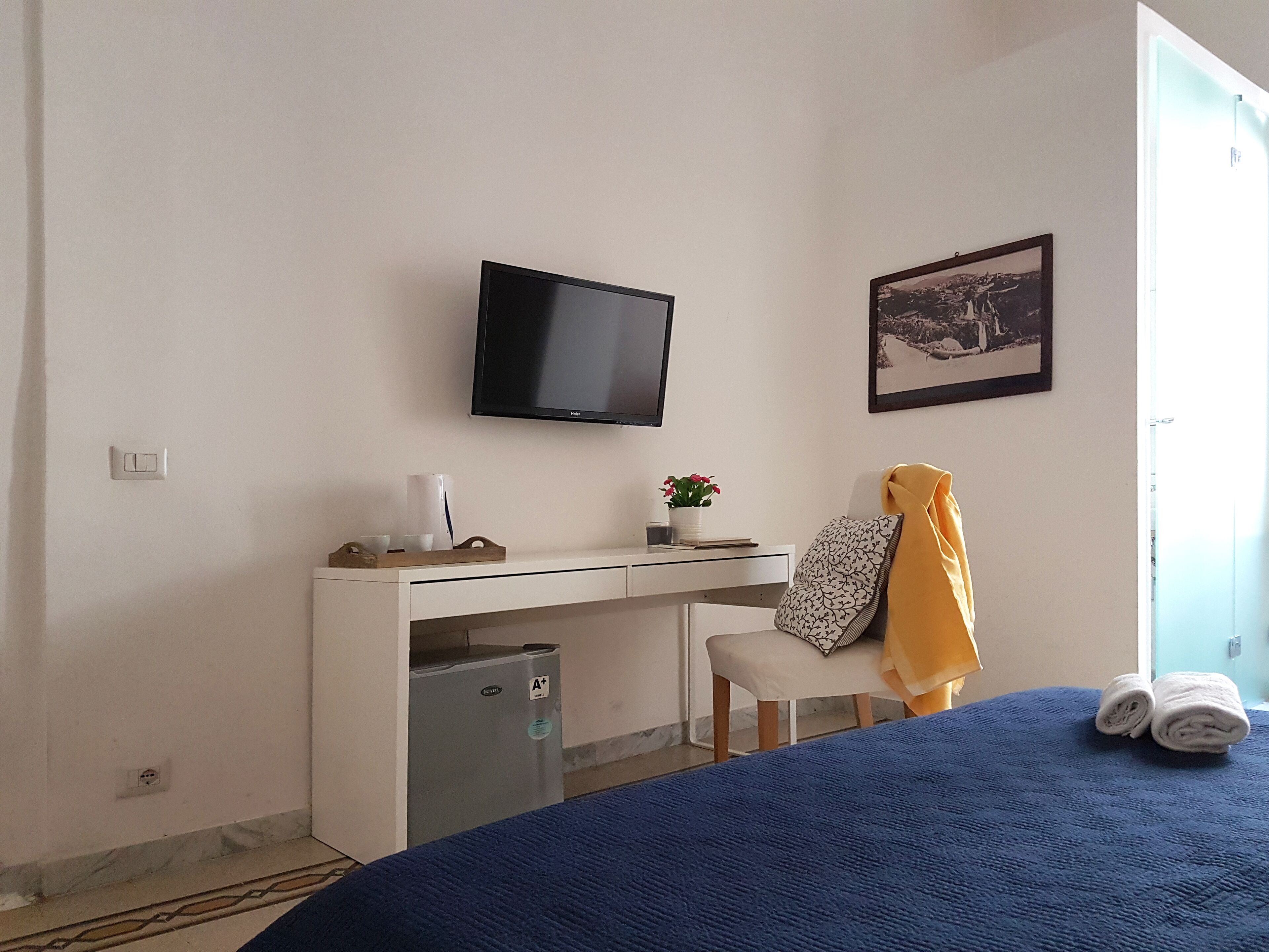 A hotel room with a bed, a desk with a computer, a television, a small fridge, a chair, and a window.