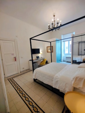Ein Schlafzimmer mit einem Himmelbett, einem Schreibtisch mit Computer, einem Sessel, einem Fernseher und einem Fenster.