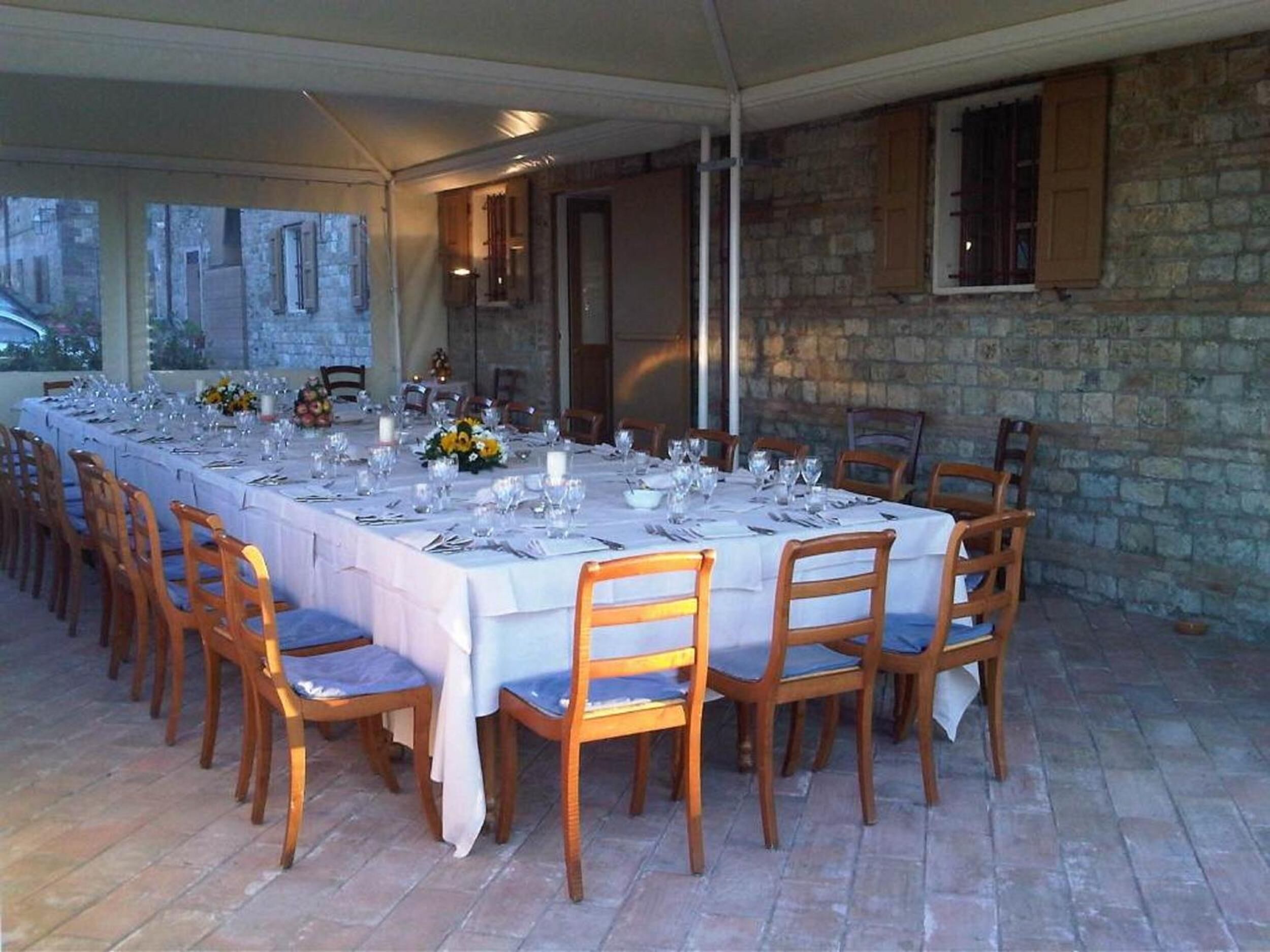 Une longue table à manger, dressée pour un repas, est installée dans un espace extérieur doté de murs en pierre et de meubles en bois.