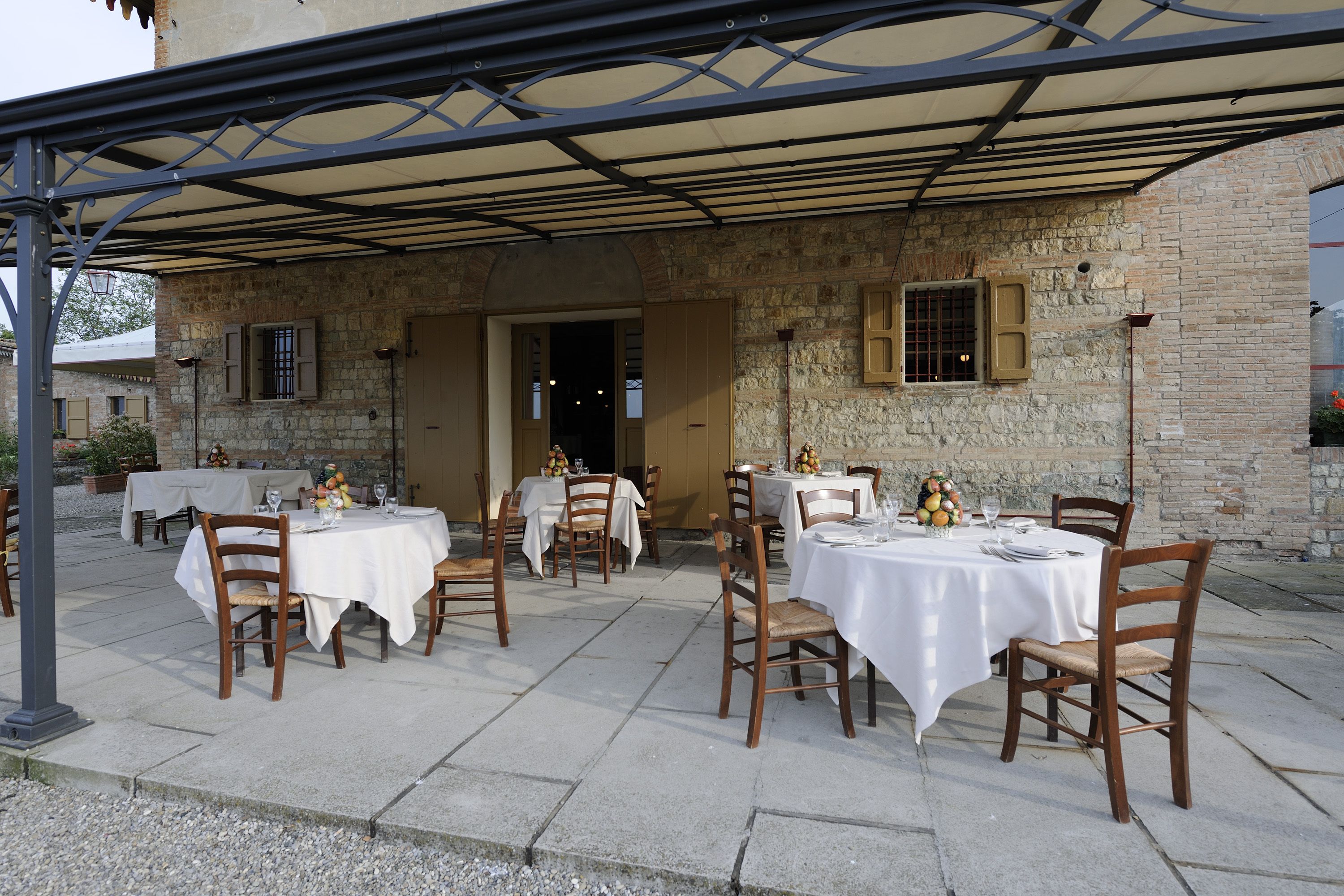 Un espace extérieur aménagé pour les repas, avec des tables et des chaises, une terrasse couverte et un bâtiment en pierre en arrière-plan.