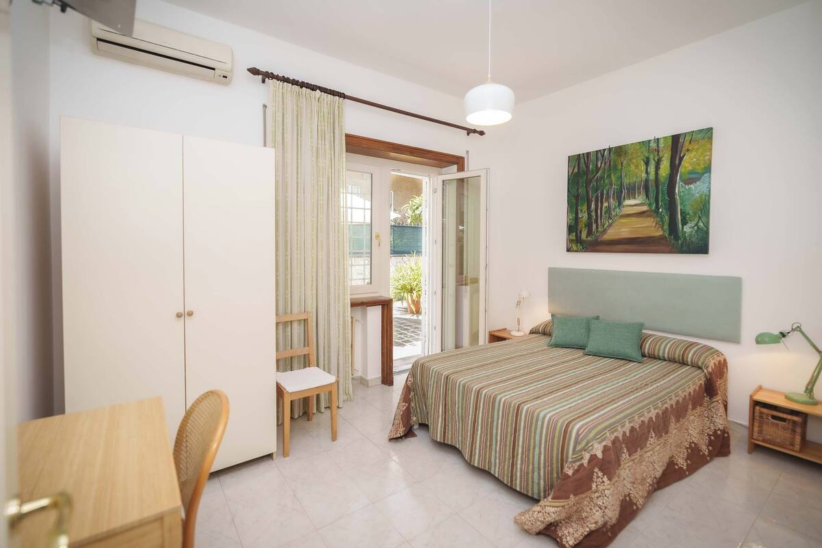 Un dormitorio con cama, un escritorio, una silla, un cuadro y una puerta que da a un balcón.