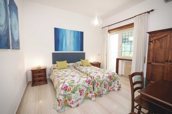 A bedroom with two beds, a wooden nightstand, a desk, and a window with curtains. - Le Tare (Rome)
