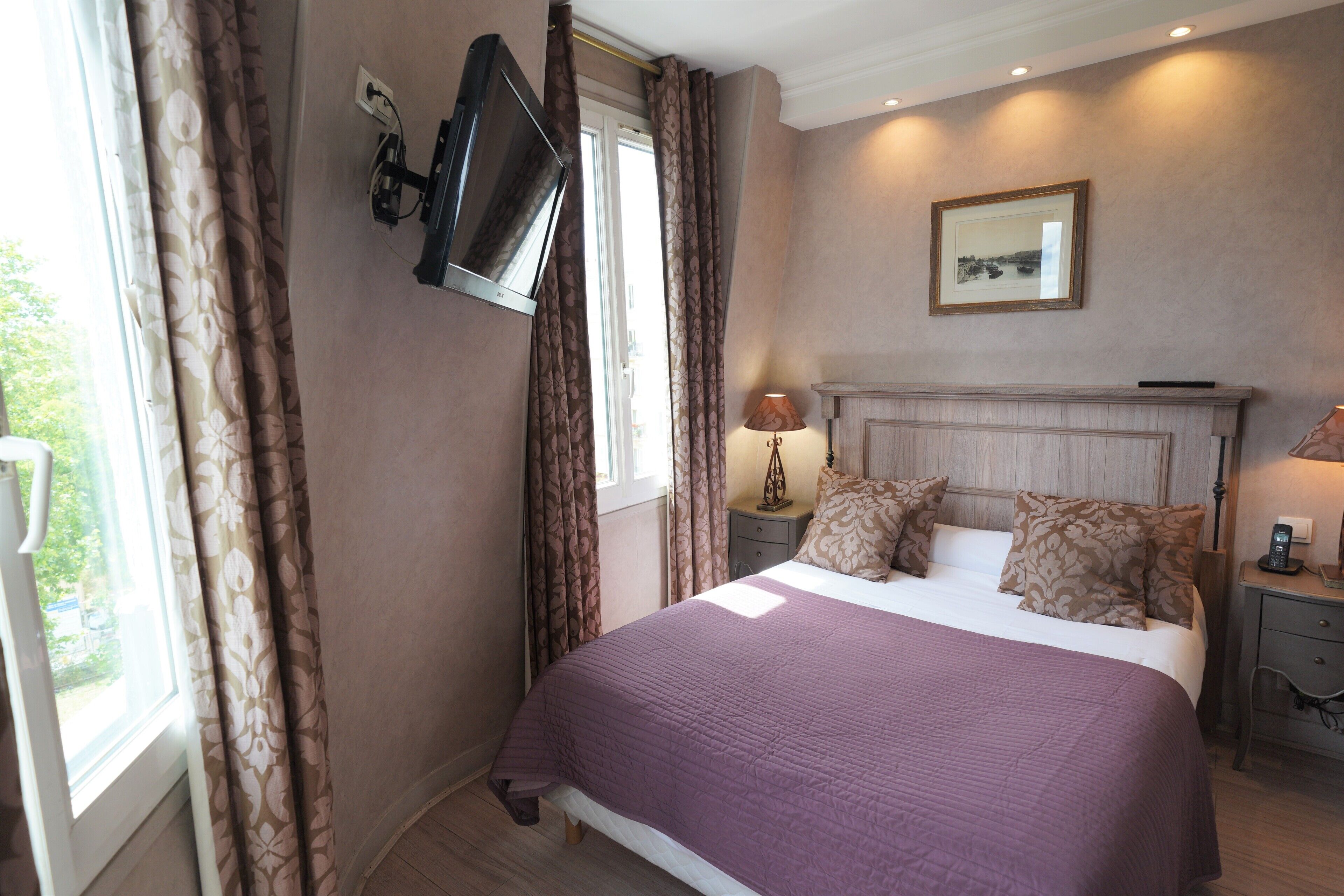 A bedroom with a bed, curtains, a TV mounted on the wall, a bedside table with a lamp, a framed picture on the wall, and a window.