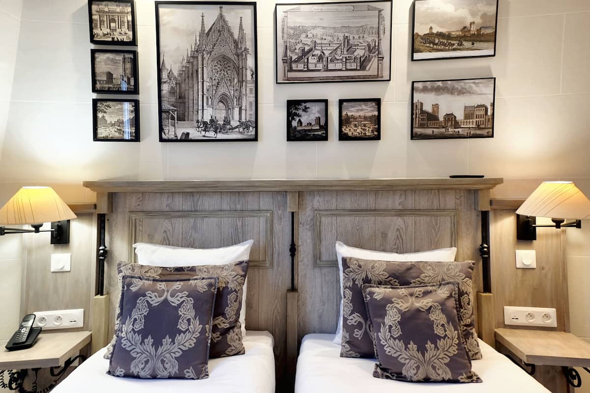 A hotel room with two beds, a wooden headboard, a nightstand with a lamp, and framed pictures on the wall.