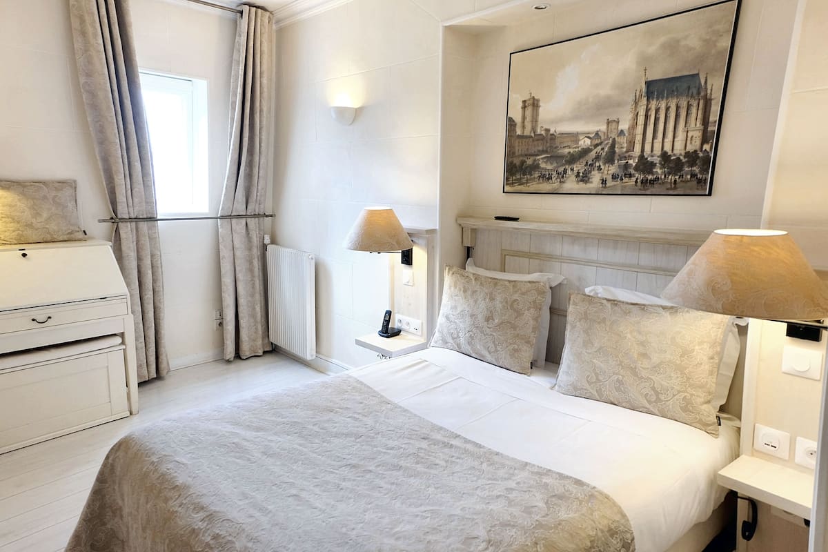 A hotel room with a bed, bedside lamps, a nightstand, a radiator, a window with curtains, and a framed cityscape painting on the wall.