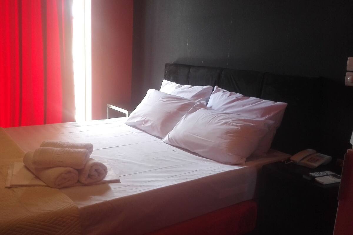 Ein Hotelzimmer mit einem Bett, Kissen, einem gefalteten Handtuch, einem Nachttischtelefon und einem Fenster mit roten Vorhängen.