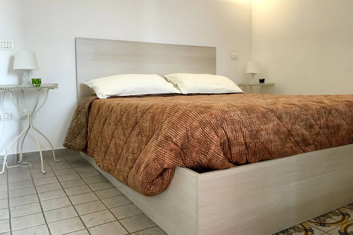 A neatly made bed with a brown quilt, a white headboard, and two bedside lamps on a white nightstand.