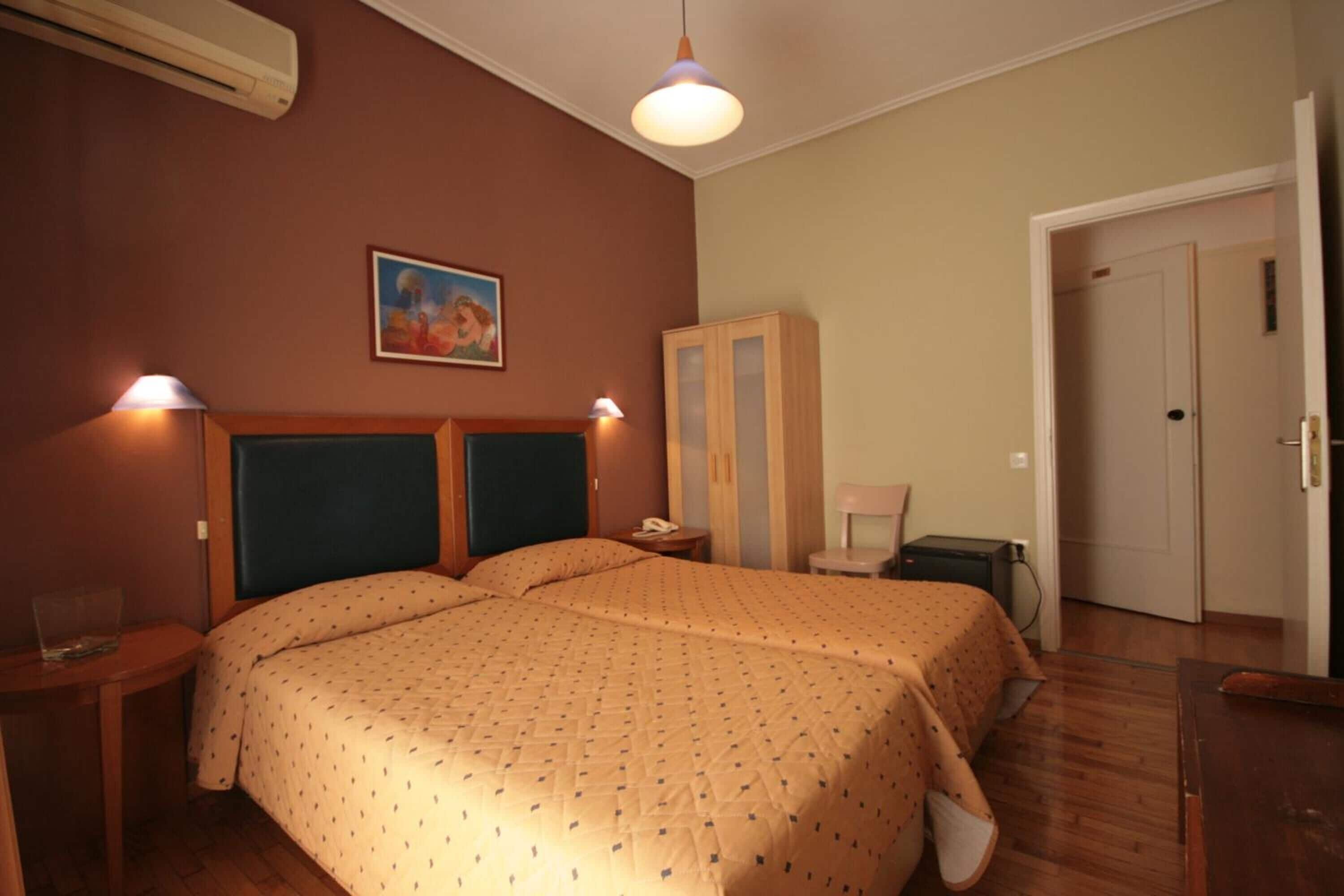 Una cama doble con una colcha estampada, una mesita de noche con una lámpara, un armario y una puerta a otra habitación.