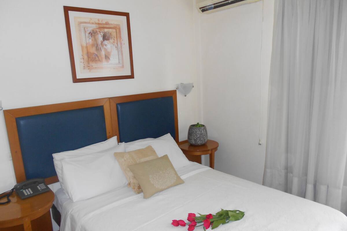 Una cama bien tendida con almohadas blancas y beige, una mesita de noche con una lámpara, un cuadro en la pared y una planta en maceta.