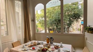 Comedor con una mesa lista para el desayuno, incluyendo platos, tazas y una cafetera. Hay sillas alrededor de la mesa y una ventana con vistas a árboles y edificios.