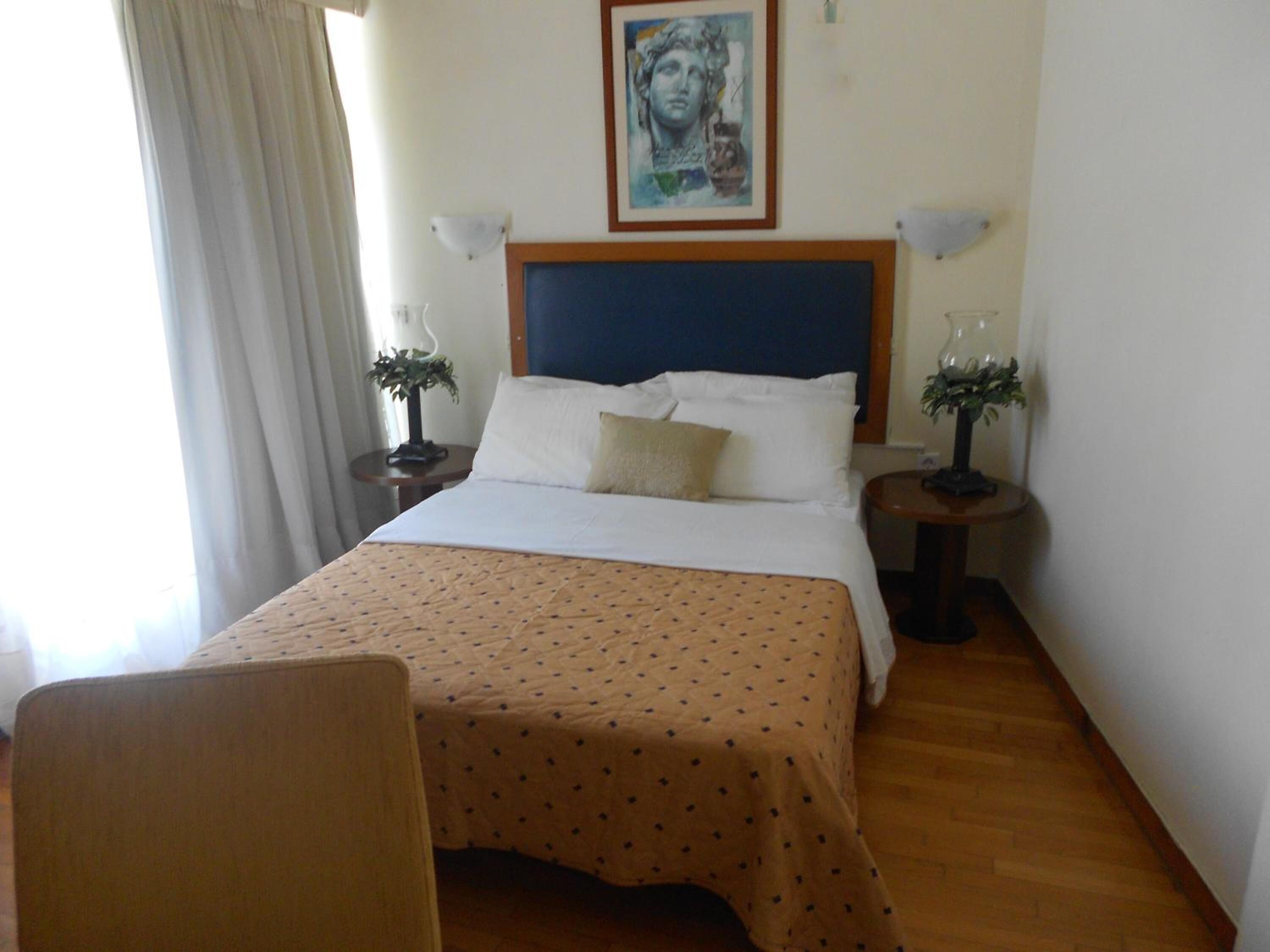 A neatly made bed with a patterned bedspread, two bedside tables with lamps, a framed artwork above the bed, and a chair in the foreground.