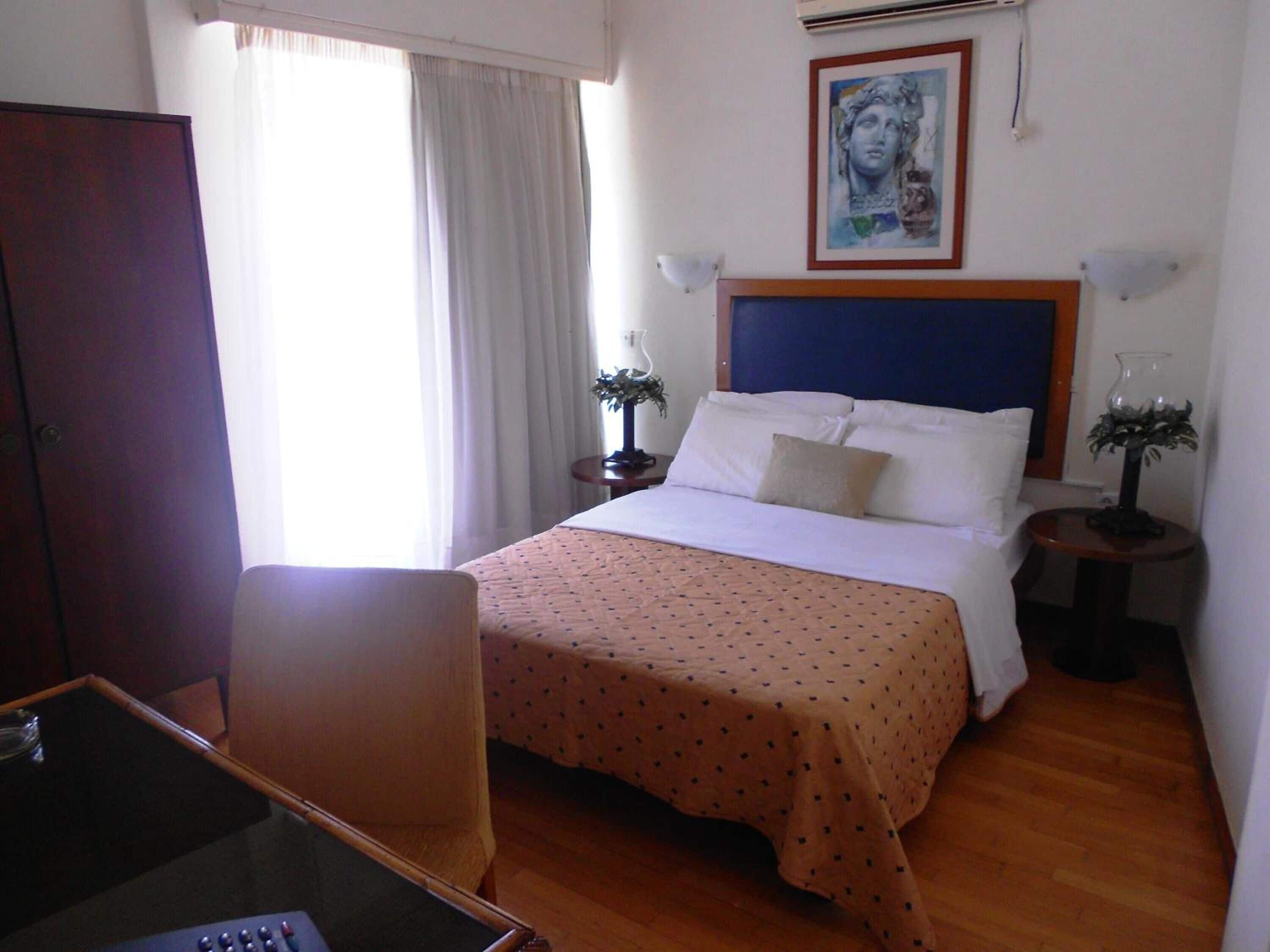 A hotel room with a bed, a desk, a chair, a window with curtains, a painting on the wall, and a nightstand with a lamp.