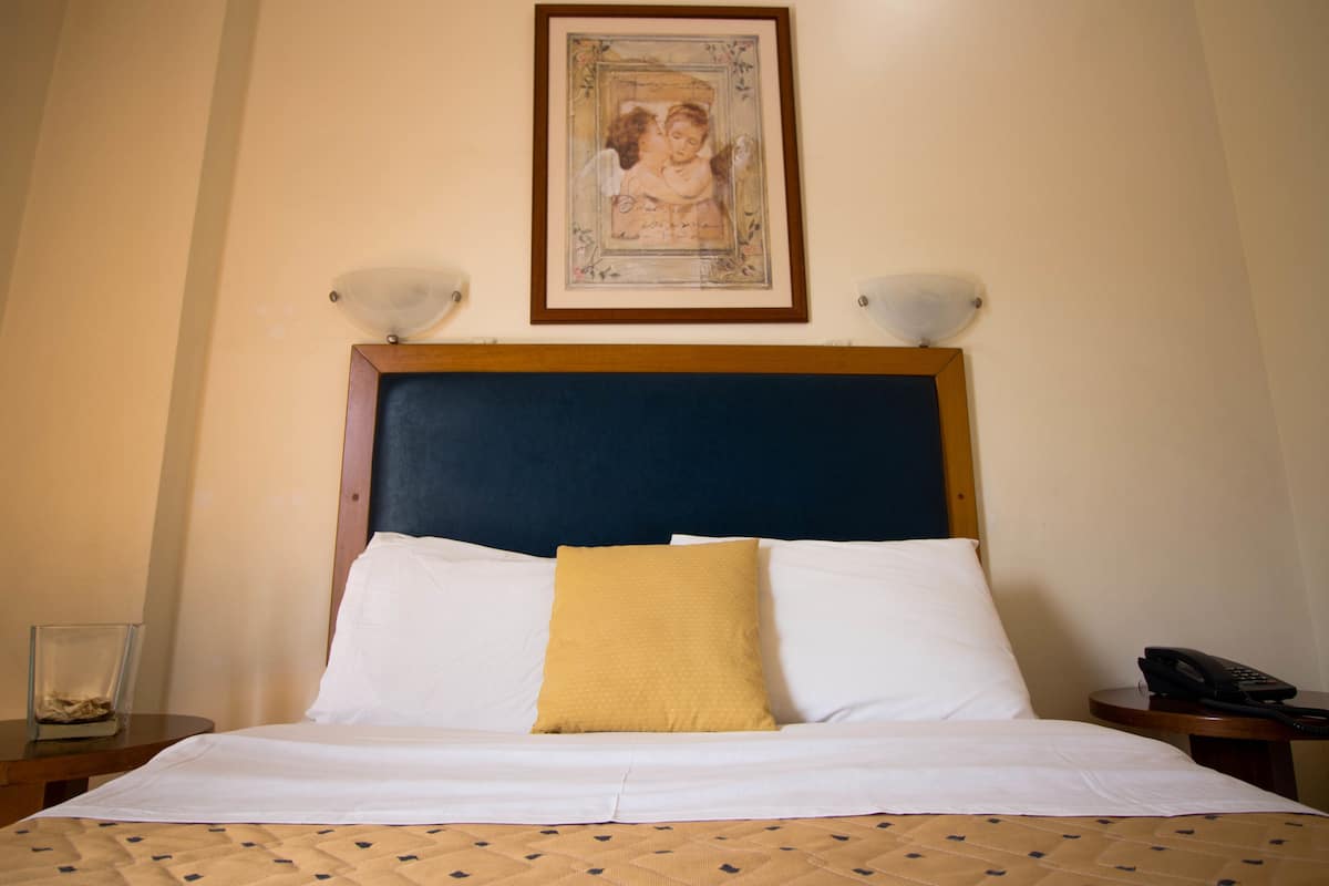 Una cama bien hecha con sábanas blancas y una almohada amarilla, un cuadro enmarcado sobre el cabecero y una mesita de noche con una lámpara y un teléfono.