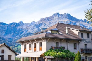 Un edificio de dos plantas con arquitectura alpina tradicional, que presenta un techo inclinado y contraventanas de madera. Al fondo, montañas y cielo despejado.