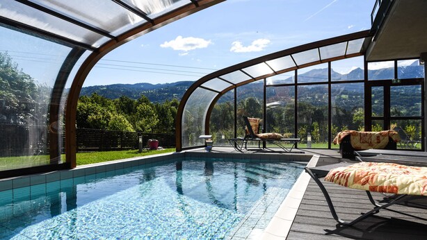 A swimming pool with a curved glass enclosure, wooden deck, and outdoor furniture. - Best Western Plus Pays du Mont Blanc (Passy)