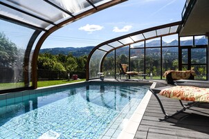 Una piscina con cerramiento de vidrio curvo, terraza de madera y mobiliario exterior.