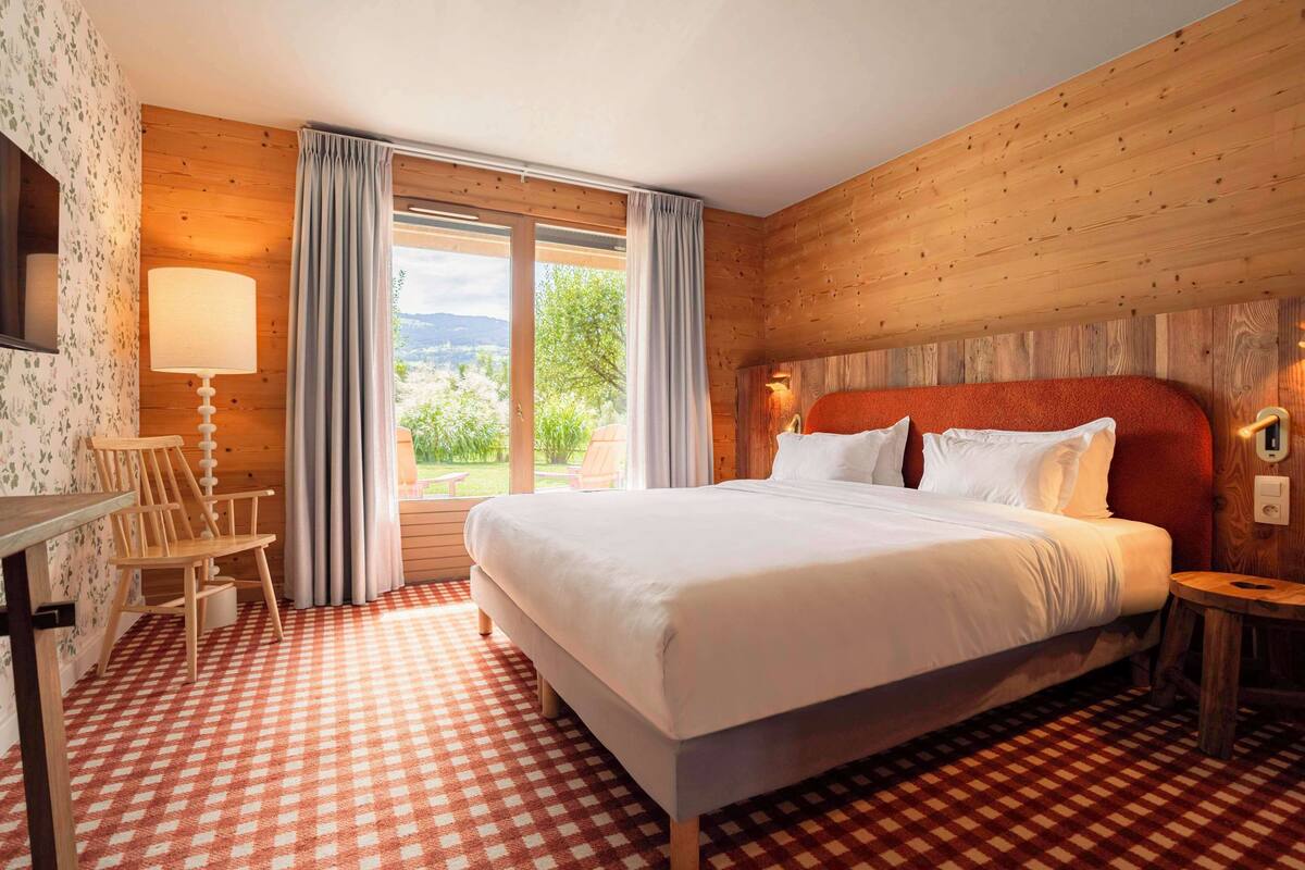 A hotel room with a large bed, wooden walls, a checkered floor, a desk with a lamp, and a view of greenery outside.