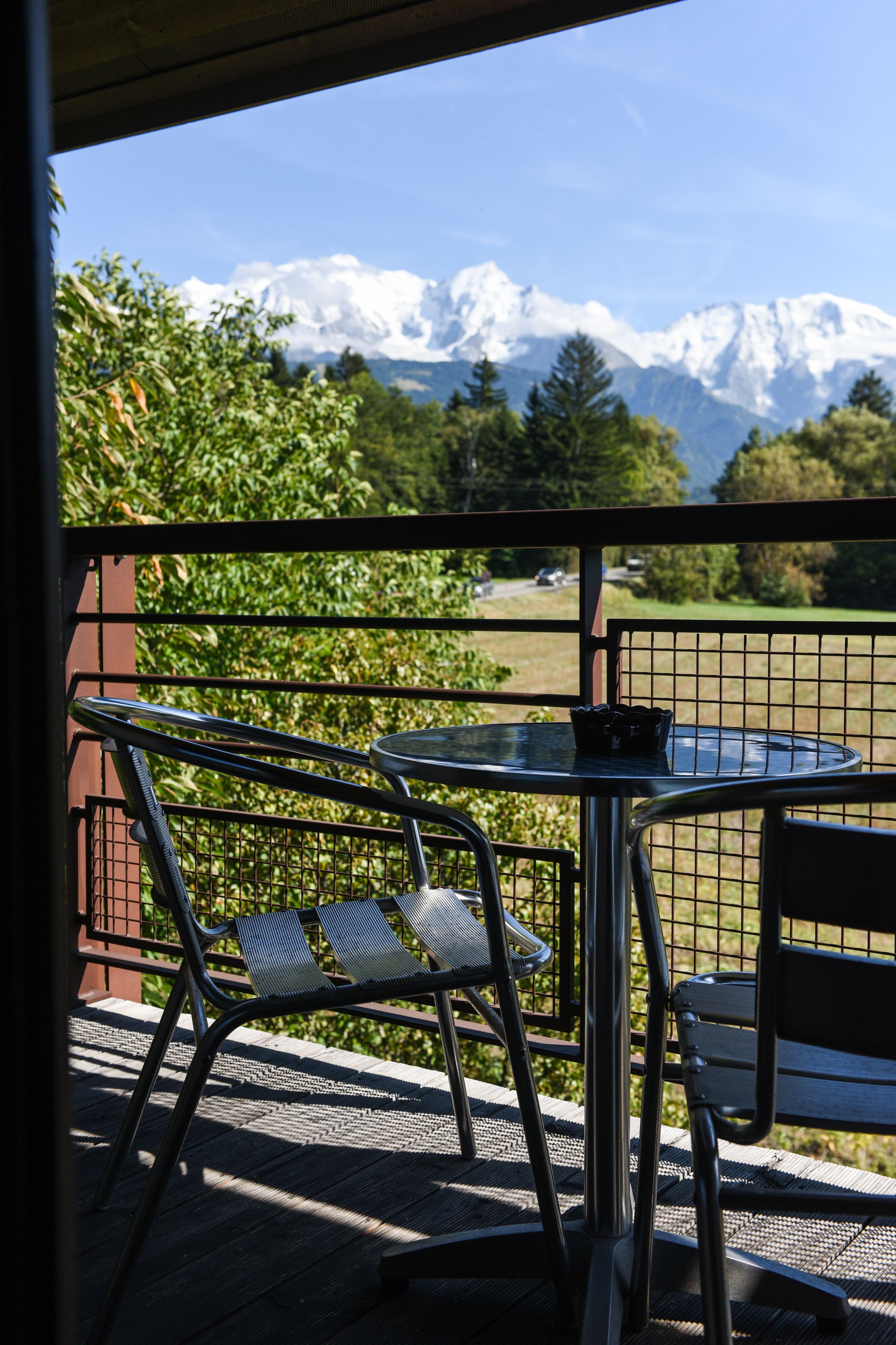 A balcony with metal chairs and a table, overlooking a mountainous landscape with snow-capped peaks.