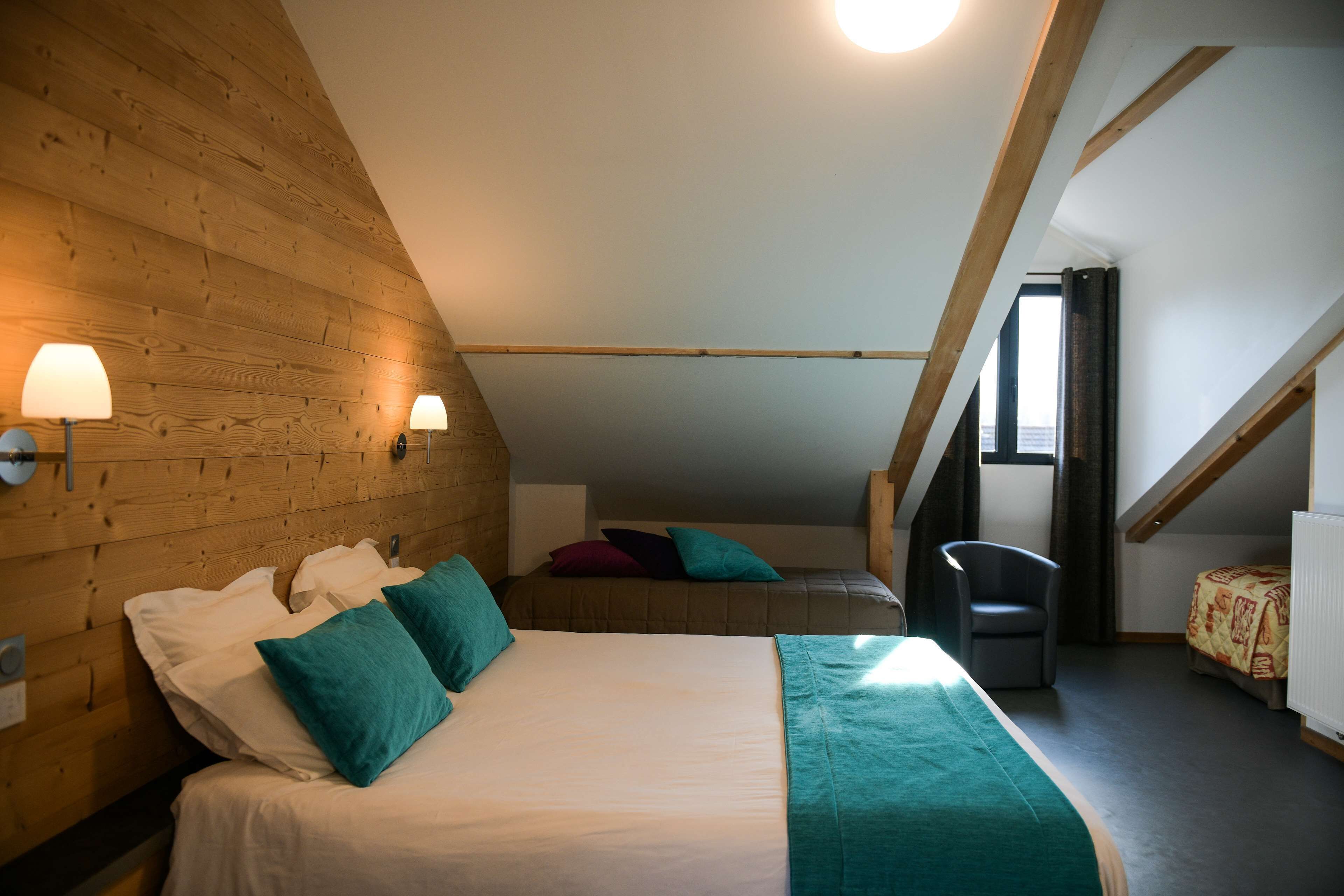 Ein Schlafzimmer mit holzverkleideter Schräge, einem Bett mit weißer Bettwäsche und Türkis-Kissen, einem Sessel und einer Heizung.