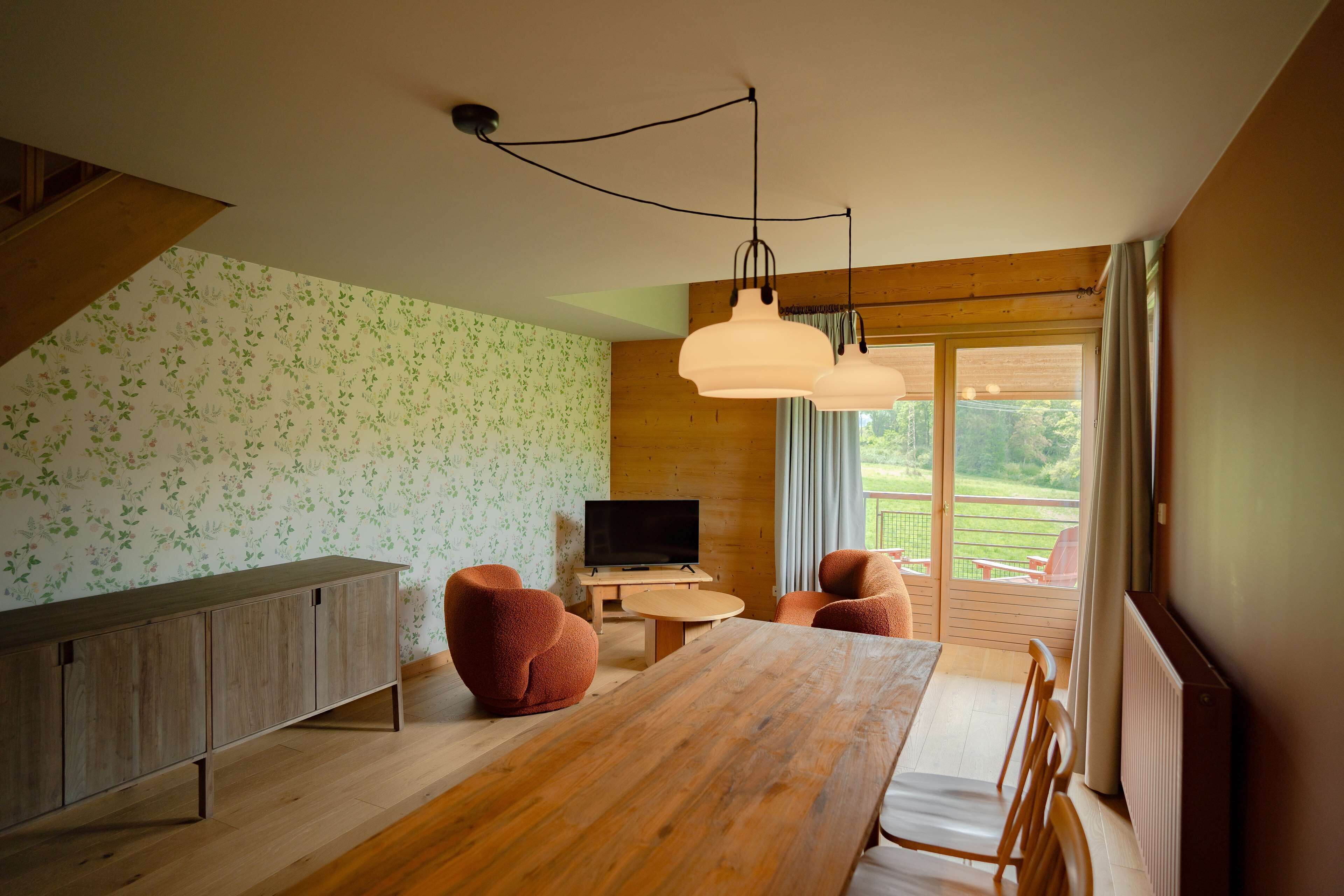 Een moderne woonkamer met een houten eettafel, twee banken, een televisie en een balkon met uitzicht op groen.