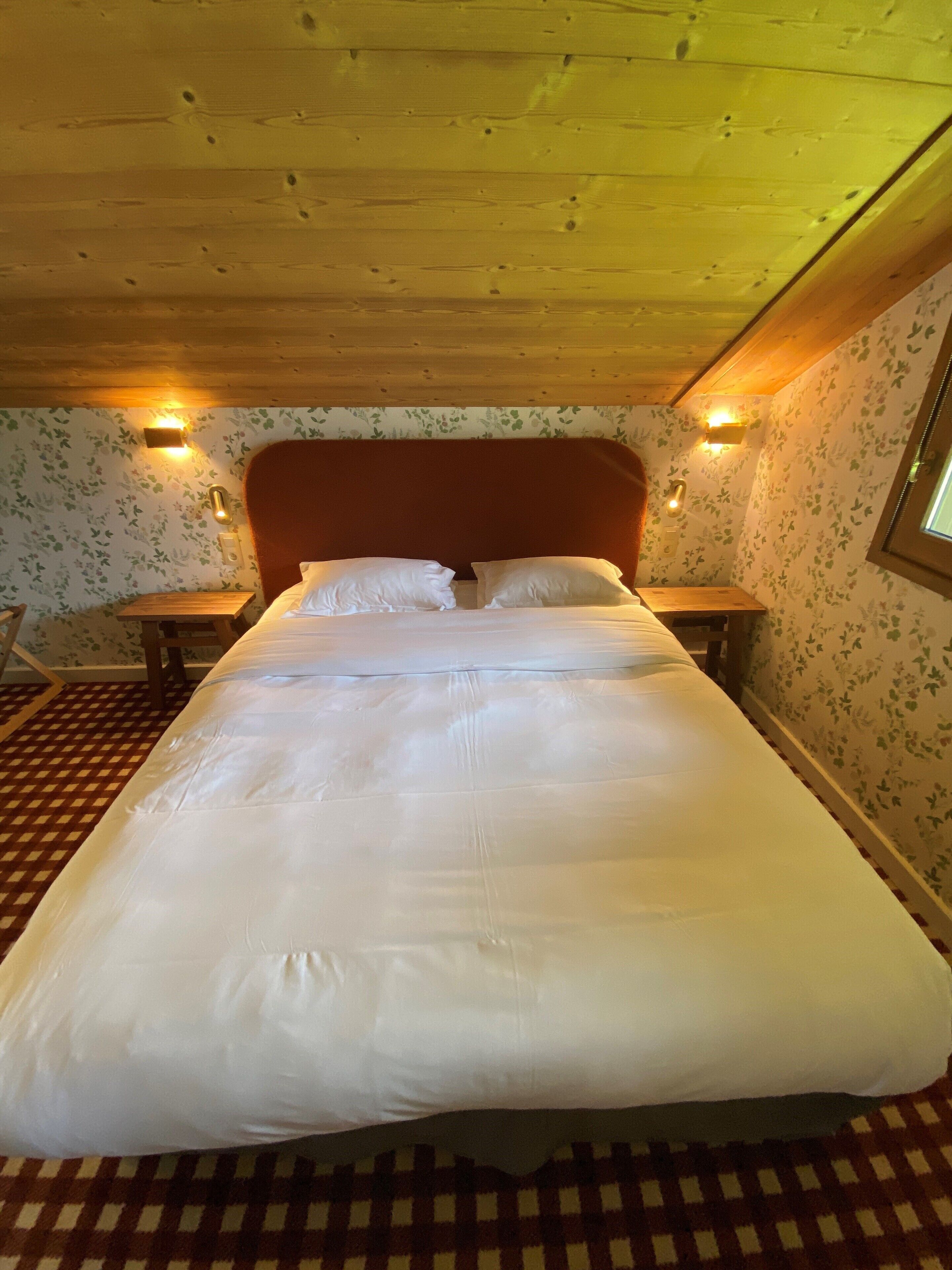 A bedroom with a large bed, wooden ceiling, floral wallpaper, and two bedside tables with lamps.