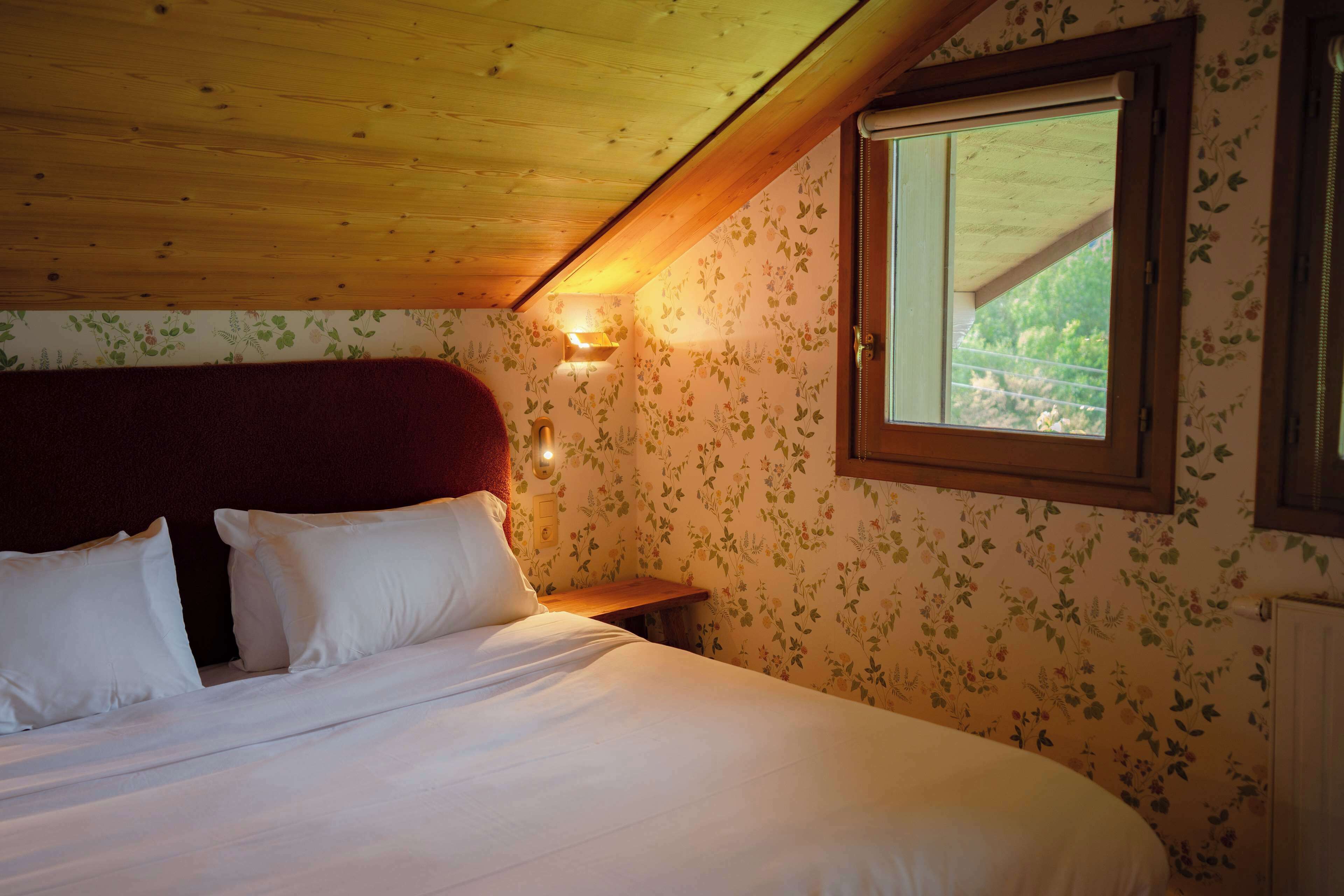 A bedroom with a wooden ceiling, floral wallpaper, a large bed with white bedding, a nightstand, and a window with a view.