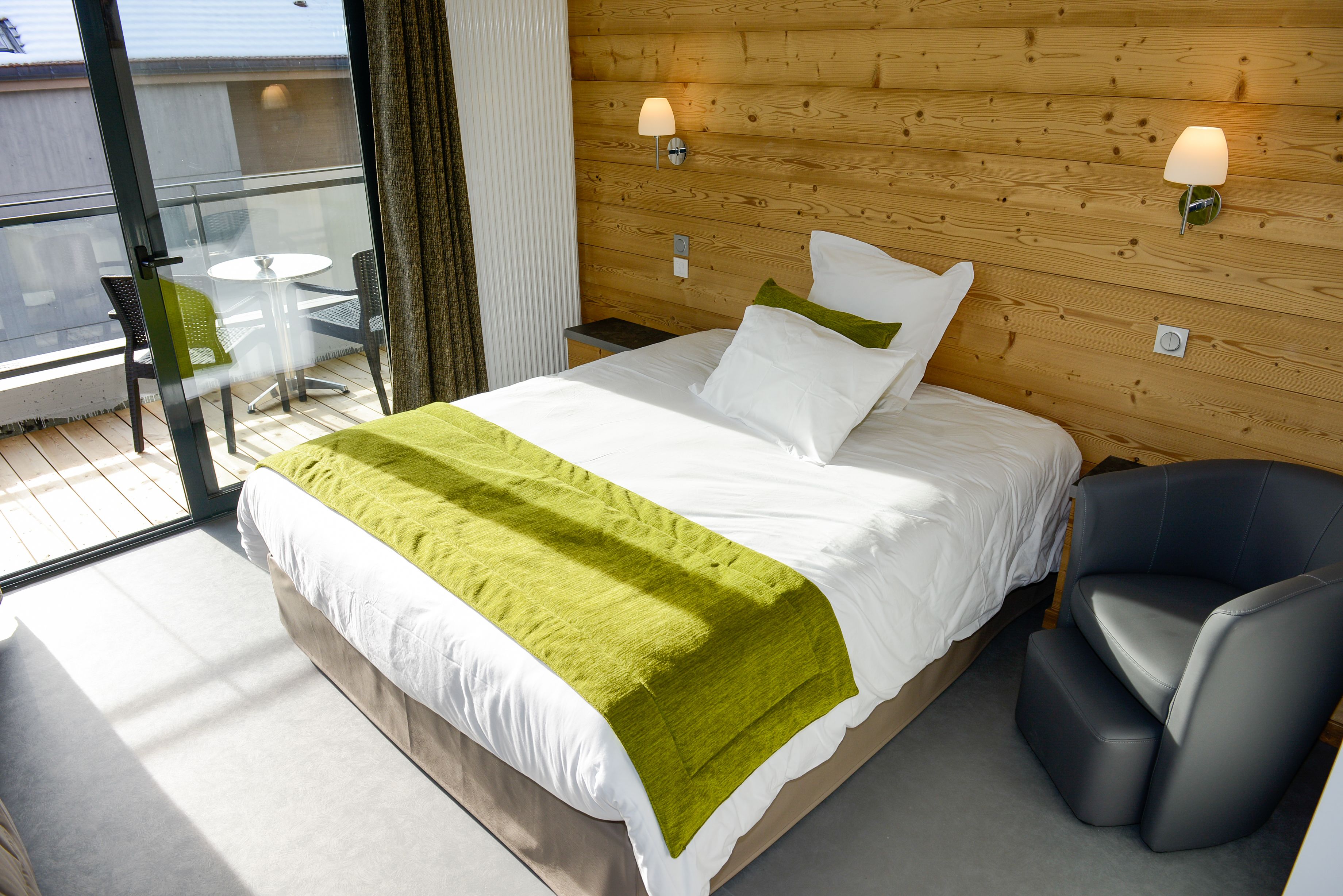 A hotel room with a bed, a green throw blanket, a nightstand, a lamp, a chair, and a view of the outside balcony.