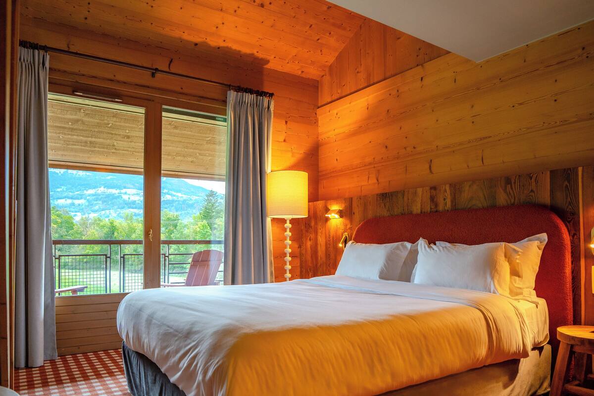 A wooden room with a large bed, checkered carpet, and a view of greenery outside.