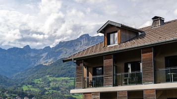 Una casa moderna de dos pisos con balcón, ventanas amplias y vista a un paisaje montañoso.