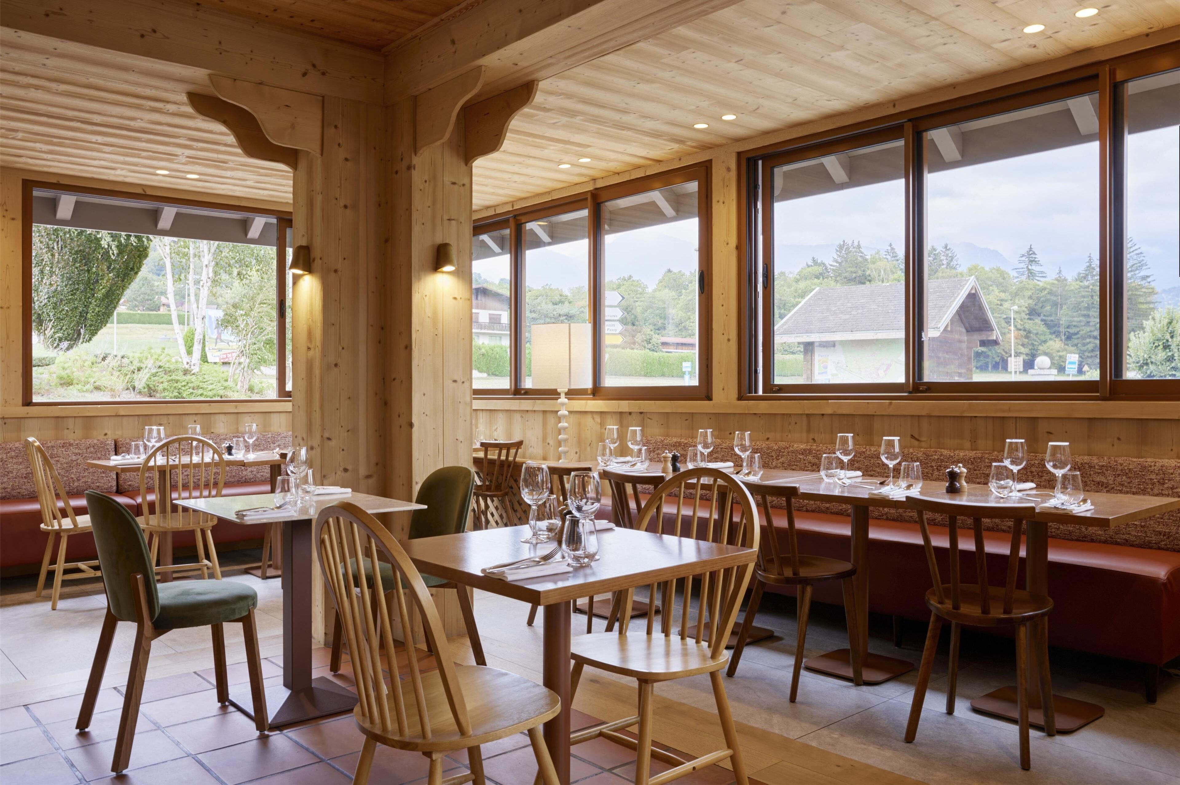 A wooden interior with tables and benches, large windows with a view of greenery and buildings.