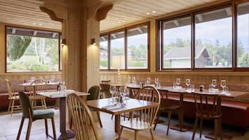 A wooden interior with tables and benches, large windows with a view of greenery and buildings.