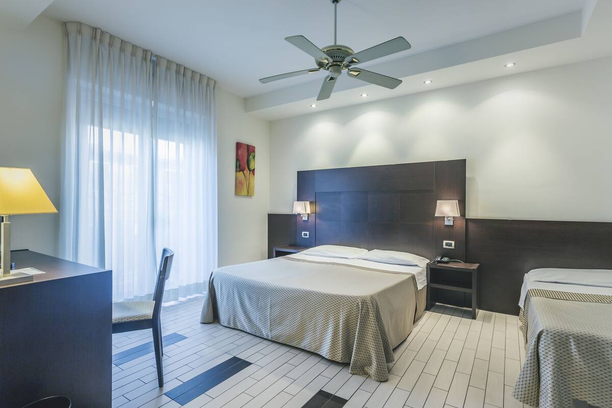 A hotel room with a large bed, a desk, a chair, a ceiling fan, and a window with curtains.