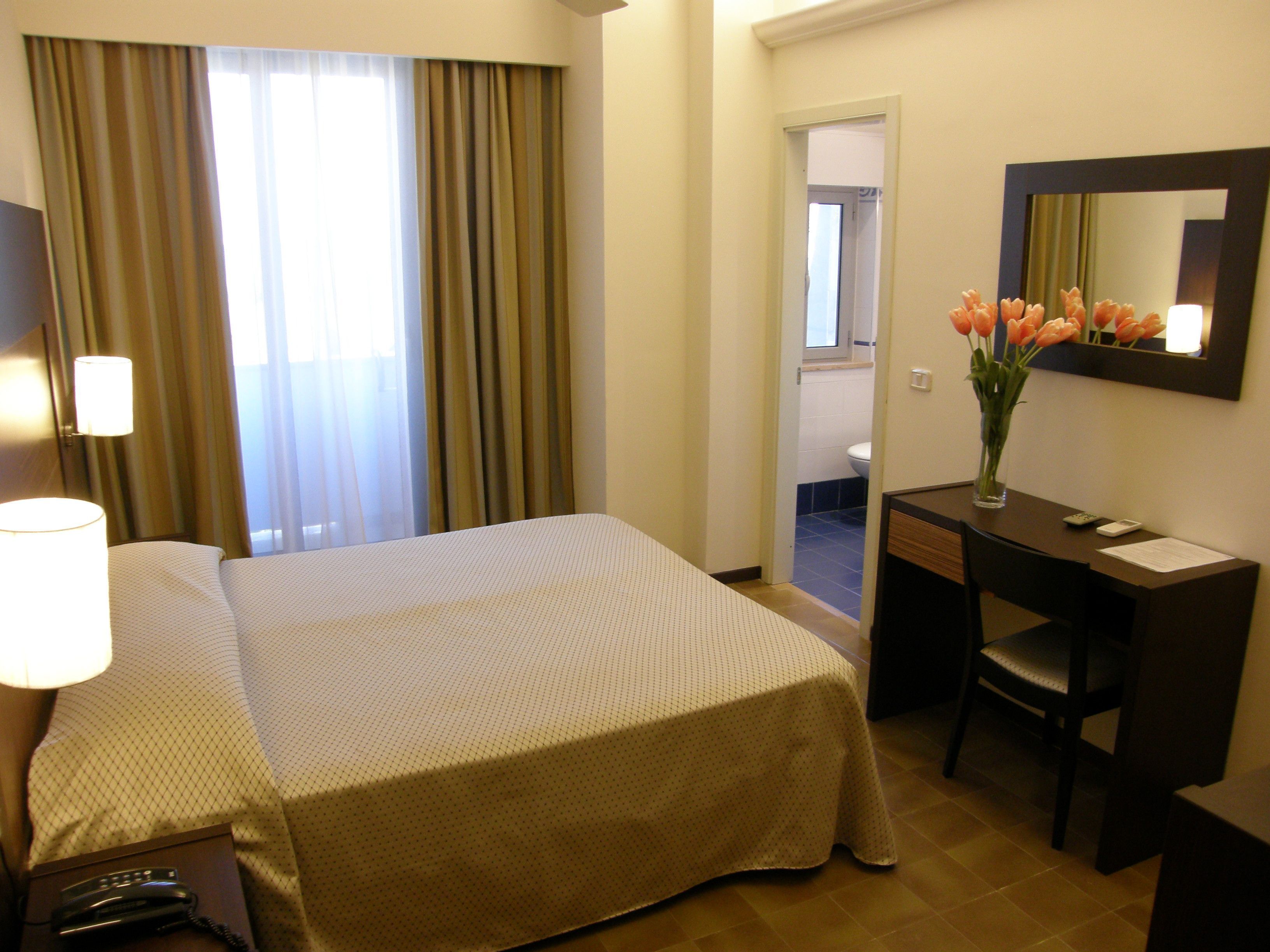 A hotel room with a bed, desk, chair, lamp, and a vase with flowers.