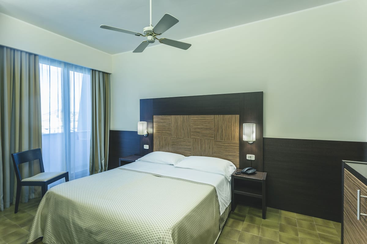 A hotel room with a bed, a chair, a desk with a lamp, a telephone, and a ceiling fan.
