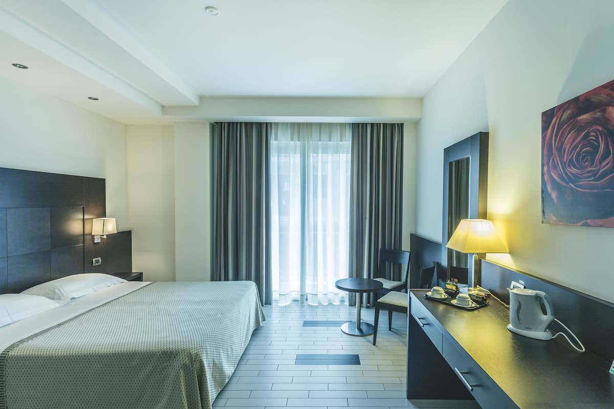 A hotel room with a bed, a desk with a lamp, a chair, a coffee maker, and a painting of a rose on the wall.