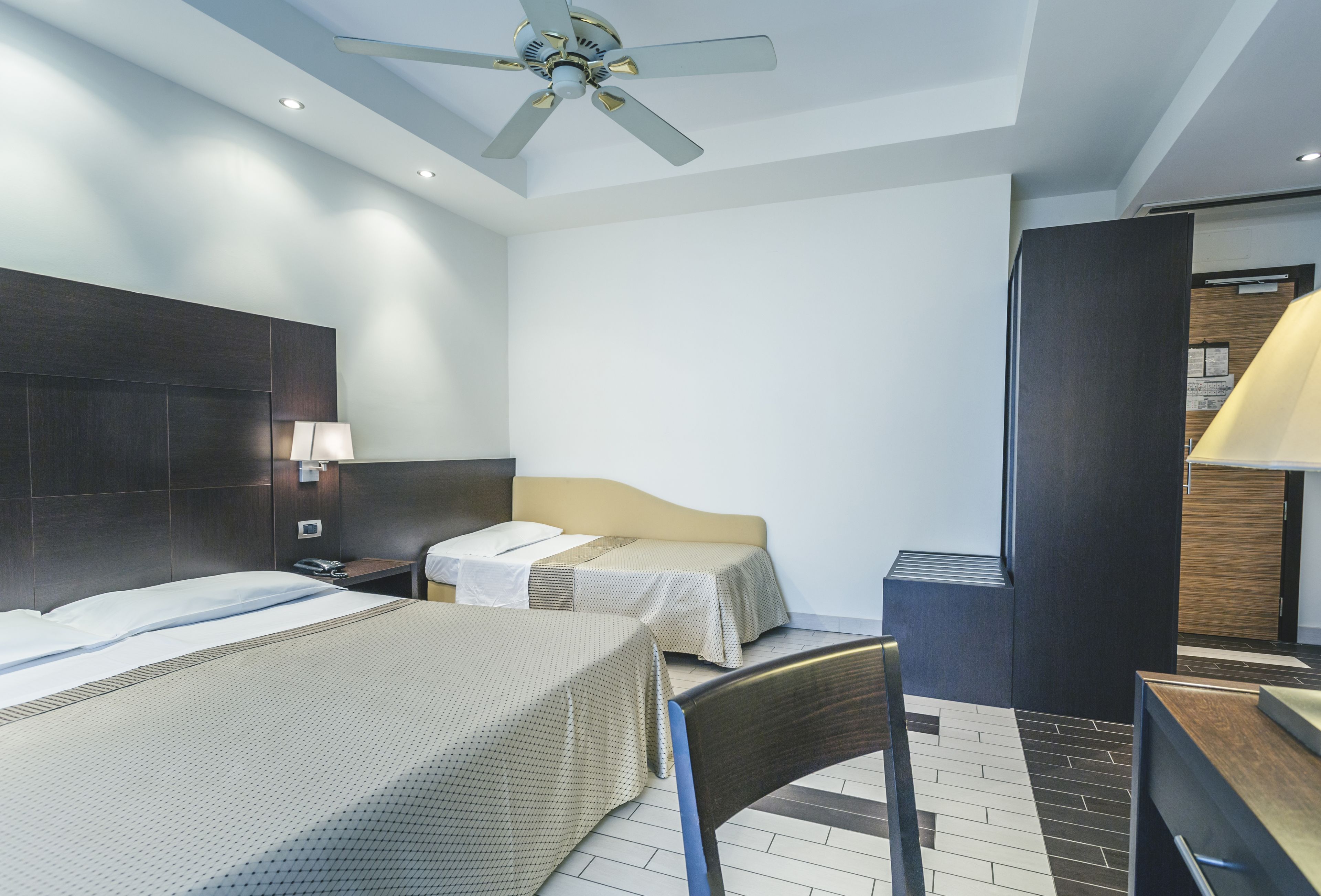 A hotel room with two beds, a ceiling fan, a desk with a lamp, and a wooden wardrobe.