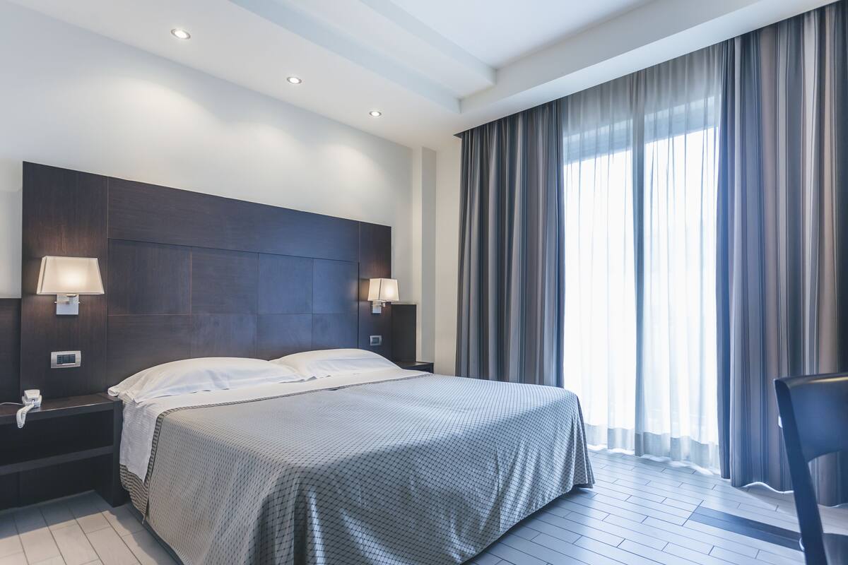 A modern hotel room with a large bed, two bedside lamps, a nightstand, a chair, and a large window with curtains.