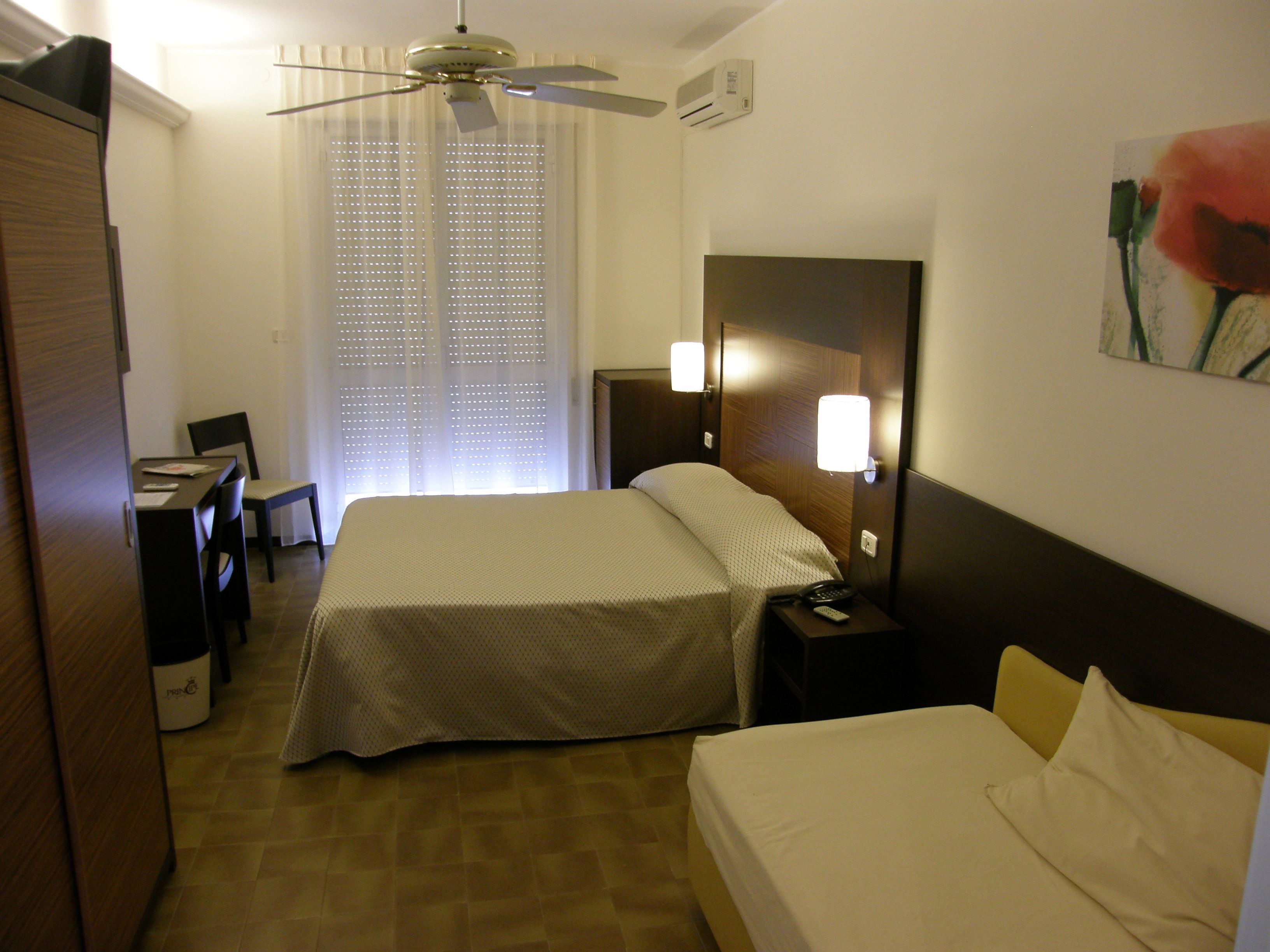 A hotel room with a bed, a desk, a chair, a nightstand, a lamp, and a ceiling fan.