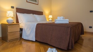 A neatly made bed with a brown bedspread, white pillows, and a white duvet. There are two bedside lamps, a wooden nightstand with a drawer, and a pair of white slippers on the floor. - Corvetto Residence (Milan)