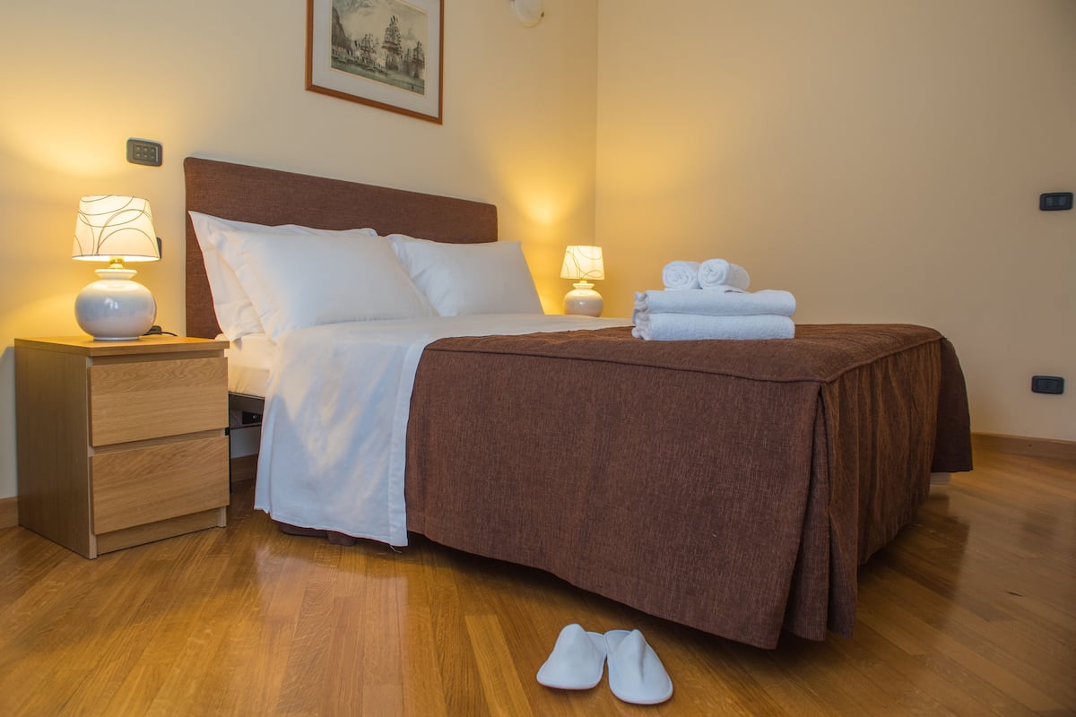 A neatly made bed with a brown bedspread, white pillows, and a white duvet. There are two bedside lamps, a wooden nightstand with a drawer, and a pair of white slippers on the floor.