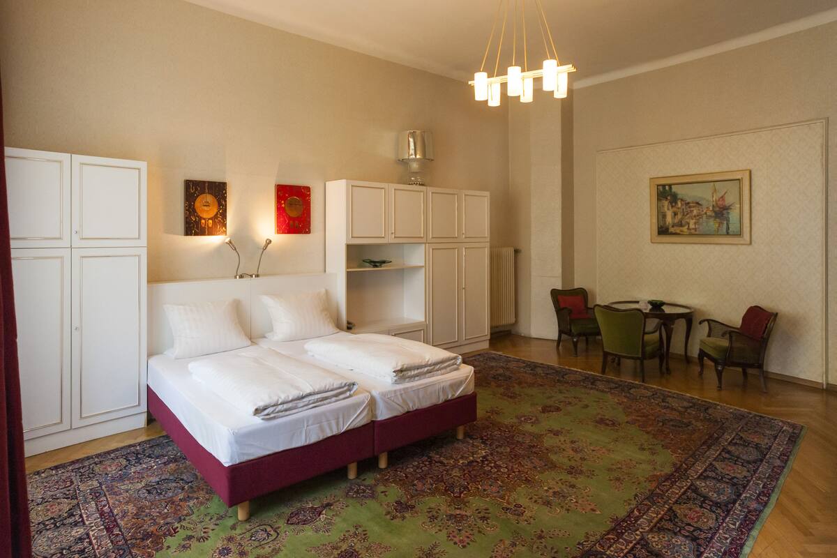 Ein Doppelbett mit rotem Kopfteil, ein weißer Kleiderschrank, ein kleiner Tisch mit Stühlen und ein Gemälde an der Wand.