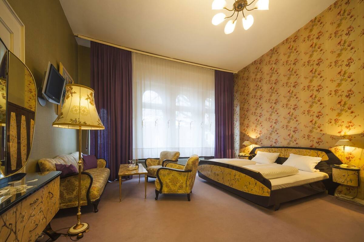 Ein Hotelzimmer mit einem Bett, zwei Sesseln, einem Sofa, einem Schreibtisch, einem Spiegel und einer Chandelier.