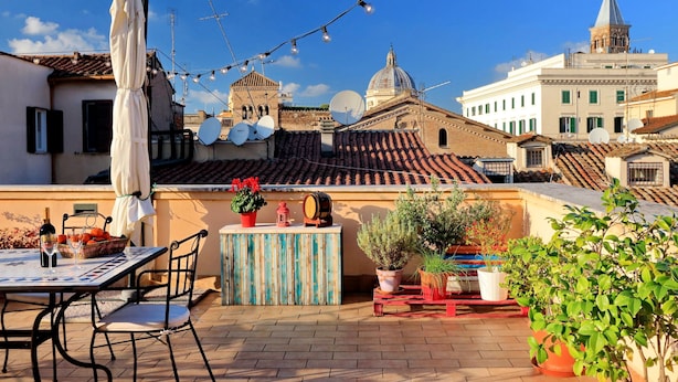 Terraza en la azotea con una mesa, sillas, plantas en macetas y una guirnalda de luces frente a un paisaje urbano.