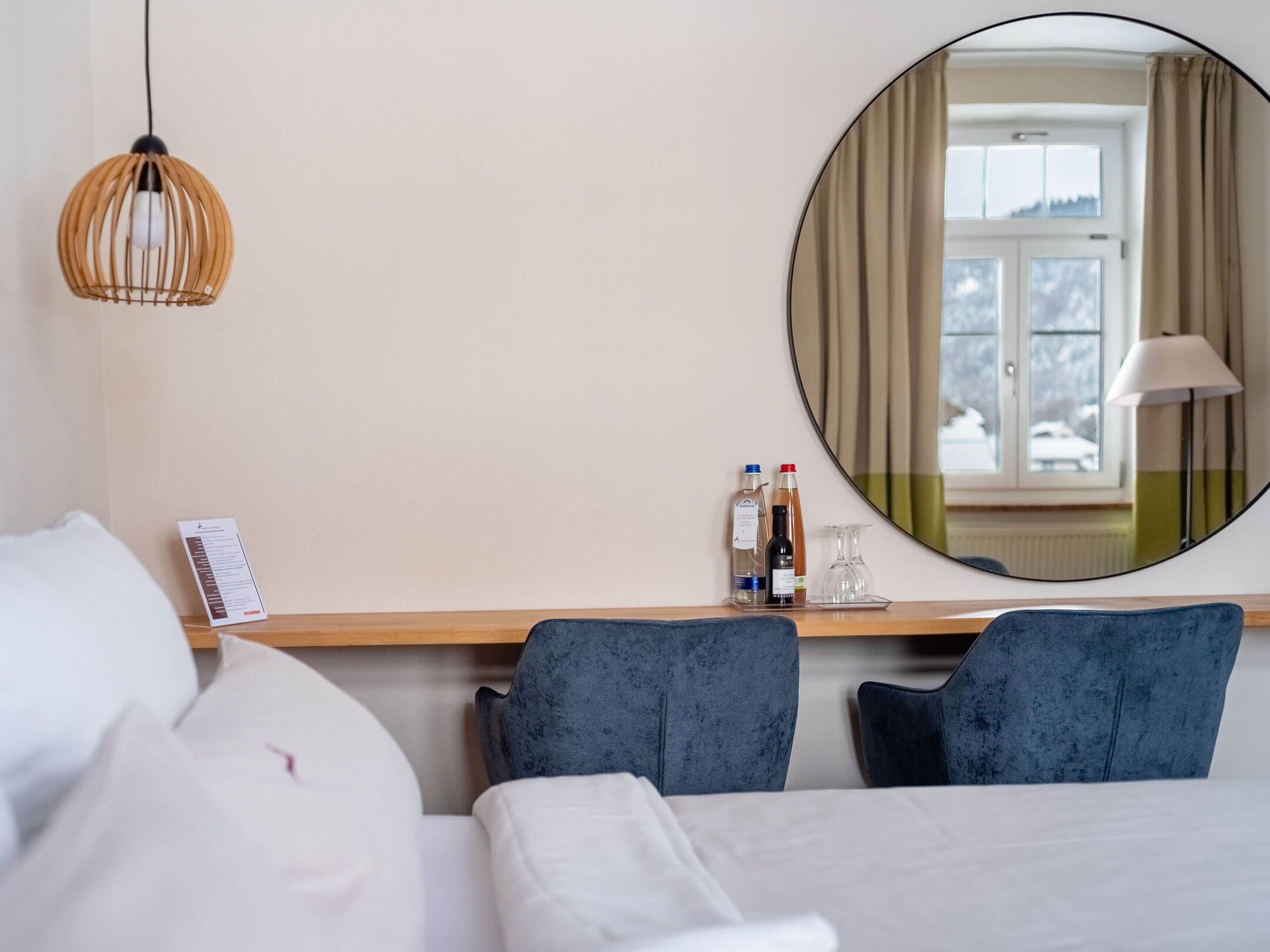 Habitación de hotel con cama, dos sillas, mesita de noche con botellas y un vaso, espejo redondo y lámpara colgante.